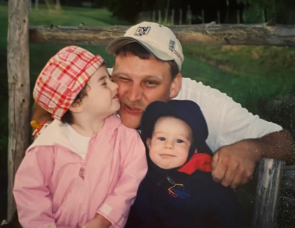 Man with two children outdoors, sitting on a log, with a wooden fence and grass in the background.